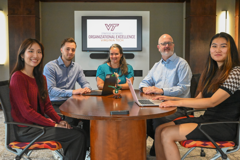 The Office for Organizational Excellence sit around a meeting table with laptops and smile for the camera.